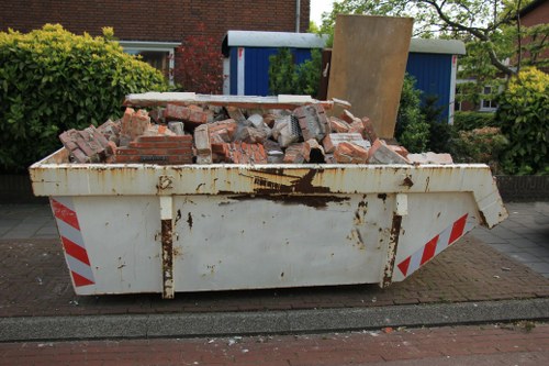 A large white metal skip bin positioned on a residential street pavement, filled with a substantial amount of broken red bricks and rubble. The bricks vary in size and appear weathered, with some dust and dirt visible on their surfaces. The skip shows signs of rust and staining, especially near the top edges, and is situated directly on a brick-paved road, with a narrow strip of concrete curb beside it. In the background, there are green shrubbery, a reddish brick building with a window, and a blue shed or garage, indicating a suburban environment. A wooden panel leans against the blue shed, and the scene is lit by natural daylight, suggesting a typical daytime setting. This image illustrates typical rubbish removal activity, possibly conducted by a company such as Commercial Waste Removal Aldgate, serving local needs for business clearance and waste disposal in the area near Aldgate.