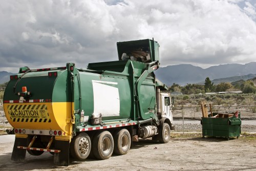 A large green commercial waste collection truck is parked on a gravel surface at an outdoor landfill or recycling site, with a partly open rear compactor indicating recent loading activity. The truck's exterior is primarily green with a yellow caution stripe at the bottom rear, and it features multiple wheels on each side for stability and weight distribution. To the right of the truck, a green wheeled waste container is visible, partially filled with various items, possibly including cardboard and mixed refuse. The surrounding environment includes a low stone wall or barrier in the background, with distant hills or mountains under a partly cloudy sky, suggesting a rural or suburban location near Aldgate or London. The scene is illuminated by natural daylight, highlighting the textures of the gravel, the metallic finish of the truck, and the varied materials in the waste container, aligning with services offered by Commercial Waste Removal Aldgate for rubbish collection and business clearance in the local area.