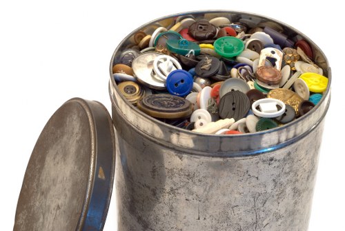 A cylindrical metal bin filled with a mixture of various plastic buttons in different sizes, shapes, and colours, including black, white, blue, green, and yellow, with some buttons partially overlapping each other. The bin shows signs of use with a slightly tarnished and scratched surface, and a loose metal lid is positioned leaning against it on the left side. The background is plain white, emphasizing the focus on the contents of the bin, which appear to be discarded small plastic items likely associated with waste collection services in Aldgate, London. The scene suggests a collection of miscellaneous small household or craft items, relevant to rubbish removal and waste management for local businesses or residents.