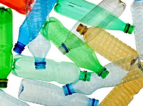 A close-up view of multiple discarded plastic bottles scattered across a flat surface, predominantly in shades of green, blue, and transparent, with some having colored caps in blue and green. The bottles vary in size and are made of smooth, glossy PET plastic, displaying typical ridged patterns near the neck and base. Some bottles are positioned horizontally, overlapping each other, with a few partially crushed or bent, indicating they are empty and used. The background is a plain white surface, providing contrast to the brightly colored bottles, emphasizing their textures and transparency. This scene highlights waste items often handled by commercial waste removal services like those offered by Commercial Waste Removal Aldgate, specializing in rubbish collection and business clearance in the Aldgate area, assisting local businesses in managing plastic waste efficiently. The environment appears clean; no other objects or environmental context are visible, focusing solely on the collection of plastic bottles for disposal or recycling purposes.