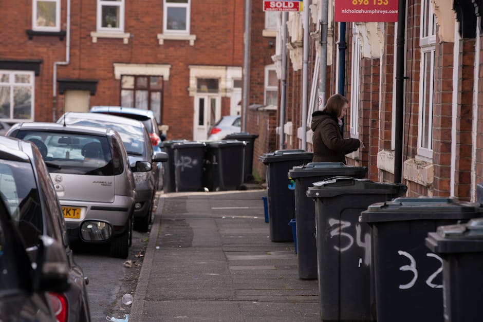 A black, traditional-style public trash bin made of metal with a rounded lid, situated on a cobblestone pavement in an urban area. The bin is overflowing with various waste items, including several cr