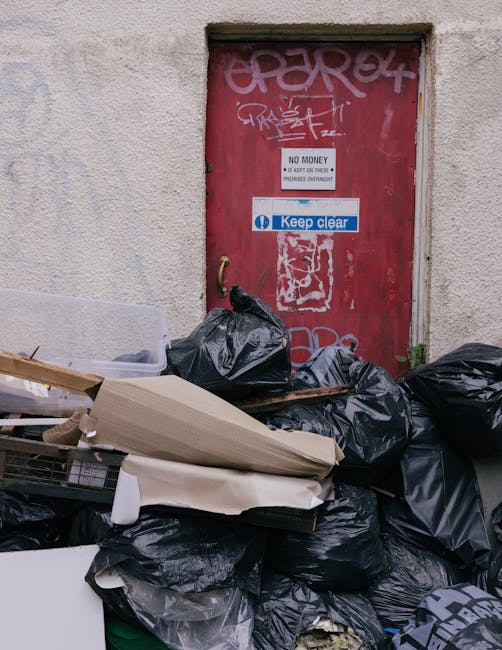 A row of several large black wheelie bins with closed lids lined up along the edge of a residential pavement on a city street, placed directly in front of brick terraced houses with white-framed windo