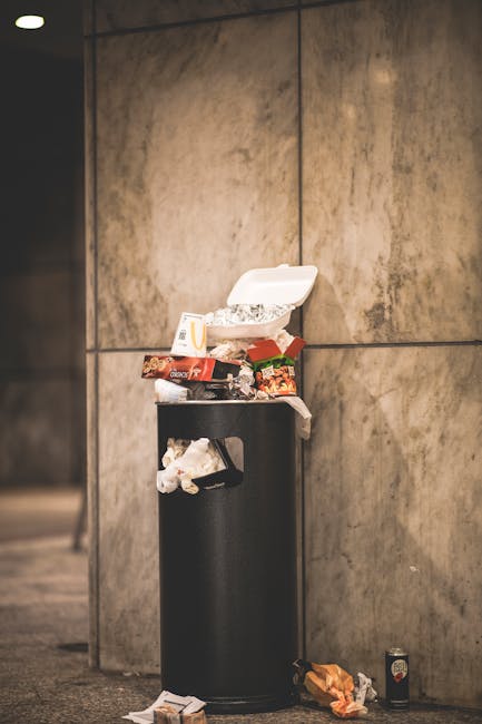 A narrow urban alleyway cluttered with a large black trash bag in the foreground, positioned on a weathered concrete surface. Behind it, a pile of various discarded waste items, including cardboard bo